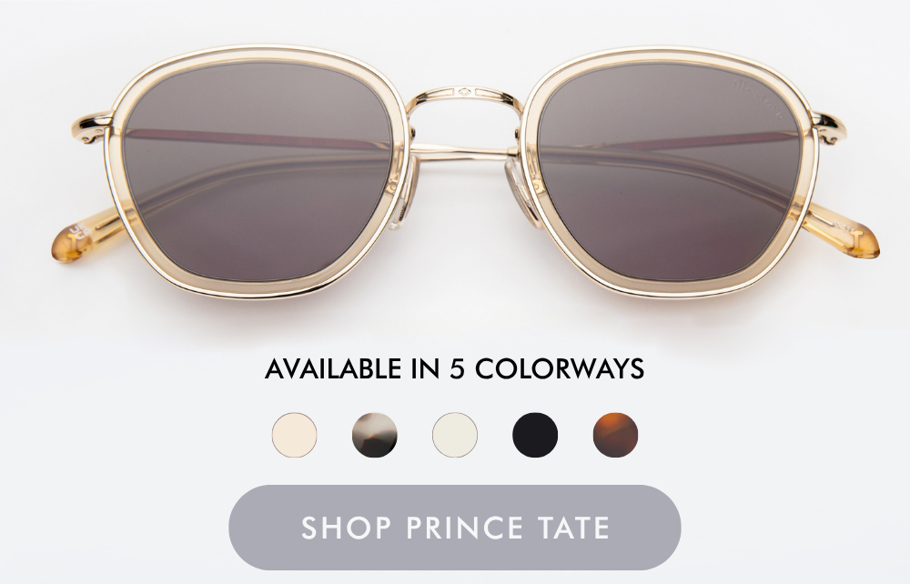 A studio product shot of the Prince Tate sunglasses, featuring a polished gold metal frame paired with clear, translucent beige acetate inner rims and dark grey lenses. Below the glasses, text reads 