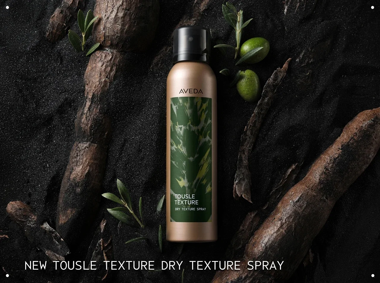 Bottle of Aveda Tousle Texture Dry Texture Spray on a black sand background with Cassava branches and green olives.