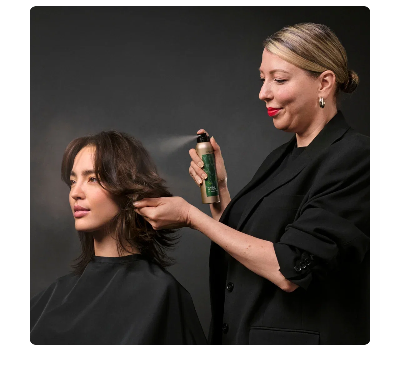Aveda stylist sprays Aveda Tousle Texture Dry Texture Spray onto a woman's brown hair.
