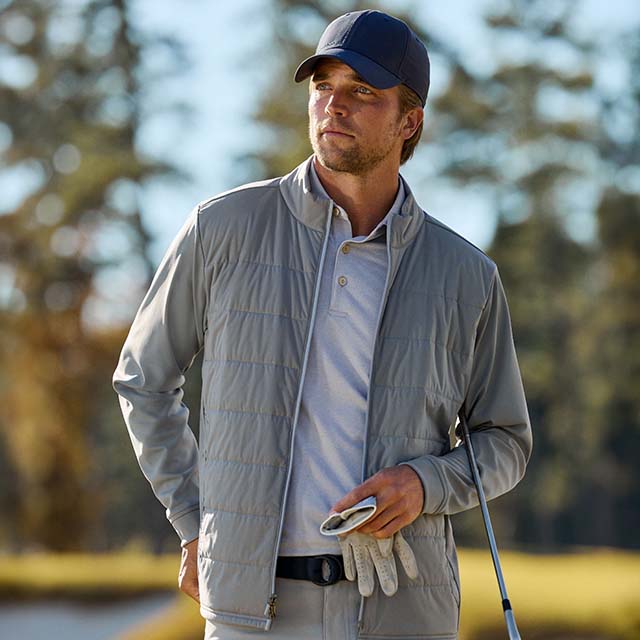 A person in golf attire, including a cap, jacket, and gloves, stands outdoors holding a golf club.