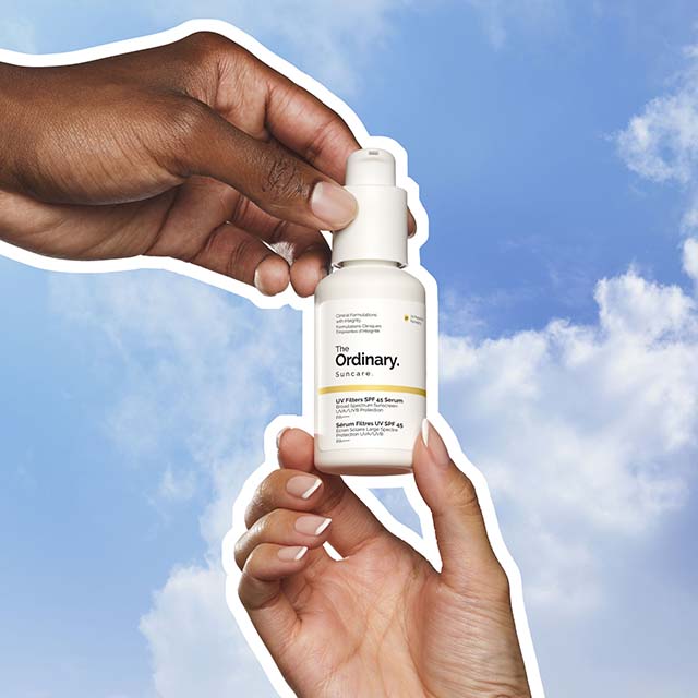 Two hands holding a bottle of The Ordinary skincare product against a blue sky.
