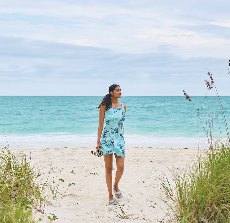 A woman walking on the beach wearing a bright blue floral sun dress and flip flops. She's carrying a fishing pole.