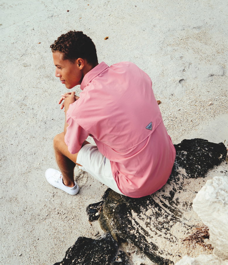 A man sitting on a rock on the beach, wearing a pink short sleeve shirt, white shorts, and sneakers.