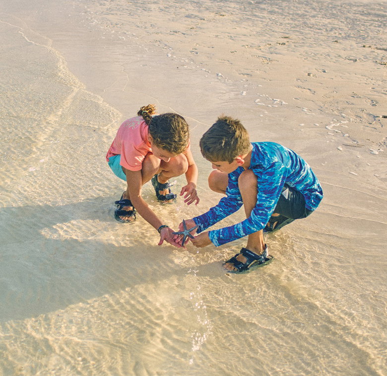 Two kids kneeling in the sand, looking at a star fish.