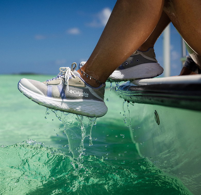 A woman hanging her legs over the side of a boat, wearing sneakers that are dripping with ocean water.