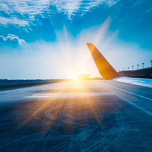 Blick über eine Start- und Landebahn am Flughafen bei Sonnenaufgang, aufgenommen aus einem Flugzeugfenster. Die Flugzeugtragfläche ist rechts im Bild zu sehen, während warmes Sonnenlicht und dramatische Wolken den Himmel erhellen.
