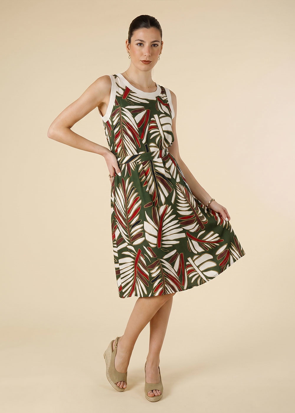 Woman modelling green tropical leaf-print sleeveless knee-length summer dress with waist tie and beige wedge sandals on neutral background
