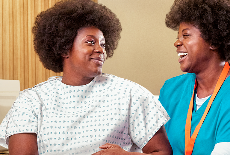 A patient wearing a patterned hospital gown sits beside a healthcare professional in blue scrubs. The nurse appears to offer support, with their arms close together in a reassuring posture. The setting suggests an indoor medical clinic or hospital examination room with warm-toned walls. The scene conveys care, trust, and compassionate medical attention.