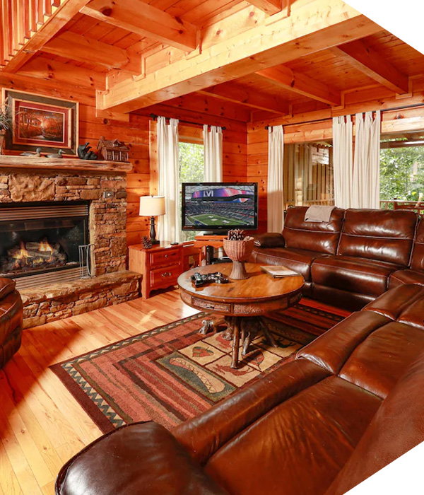 Rustic log cabin family room with leather sectional, stone fireplace, wood beams, round coffee table, and TV.