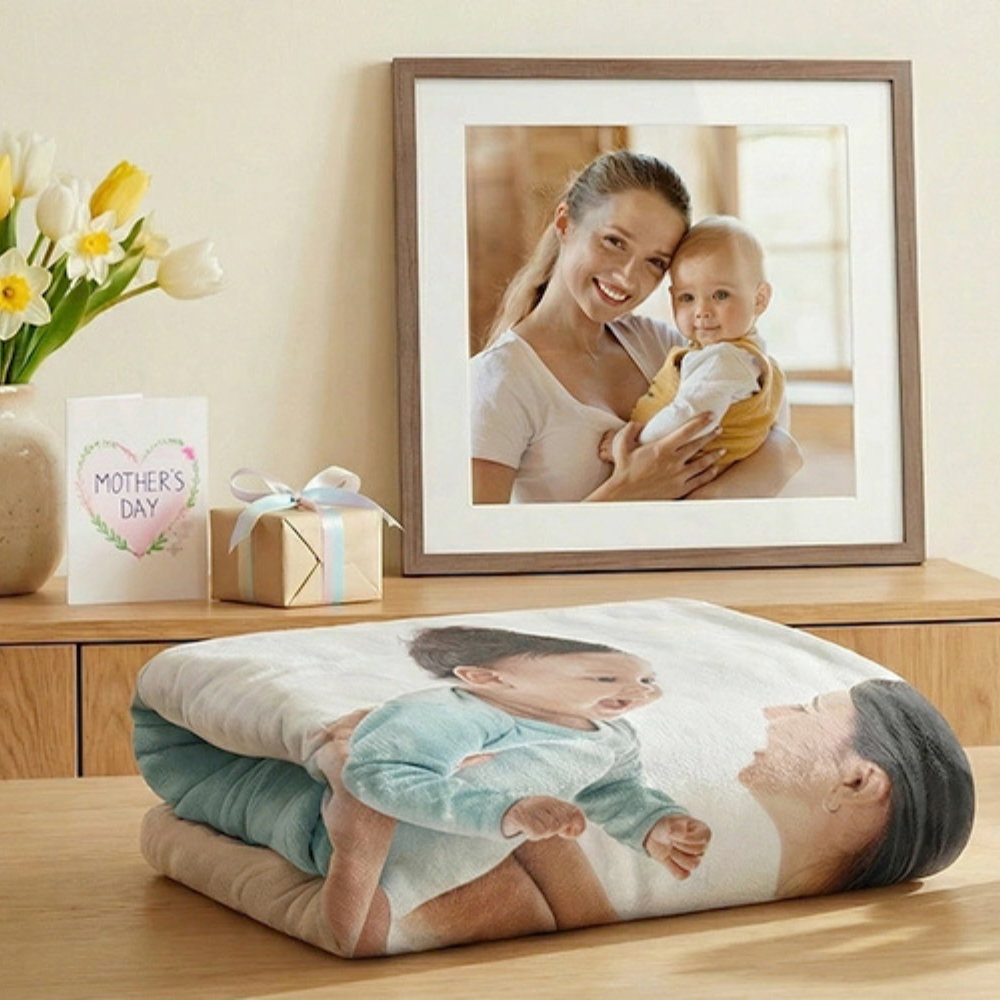 Gefaltete Decke mit Foto eines Babys und einer Frau auf einem Holztisch vor einem gerahmten Foto einer Frau, die ein Baby hält, neben einer Muttertagskarte und einem Geschenk