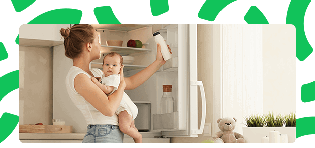 A woman holding her baby and getting a bottle of milk out of the fridge