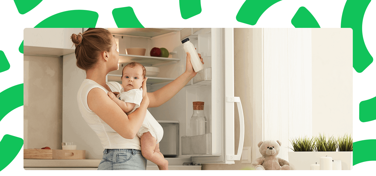 A woman holding her baby and getting a bottle of milk out of the fridge