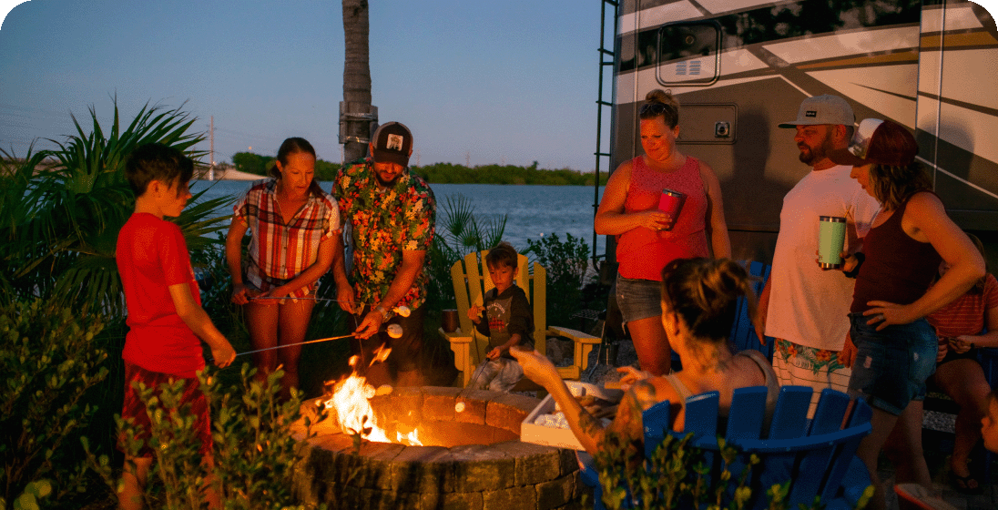 A large group roasting marshmallows over a campfire outside of an RV at dusk.