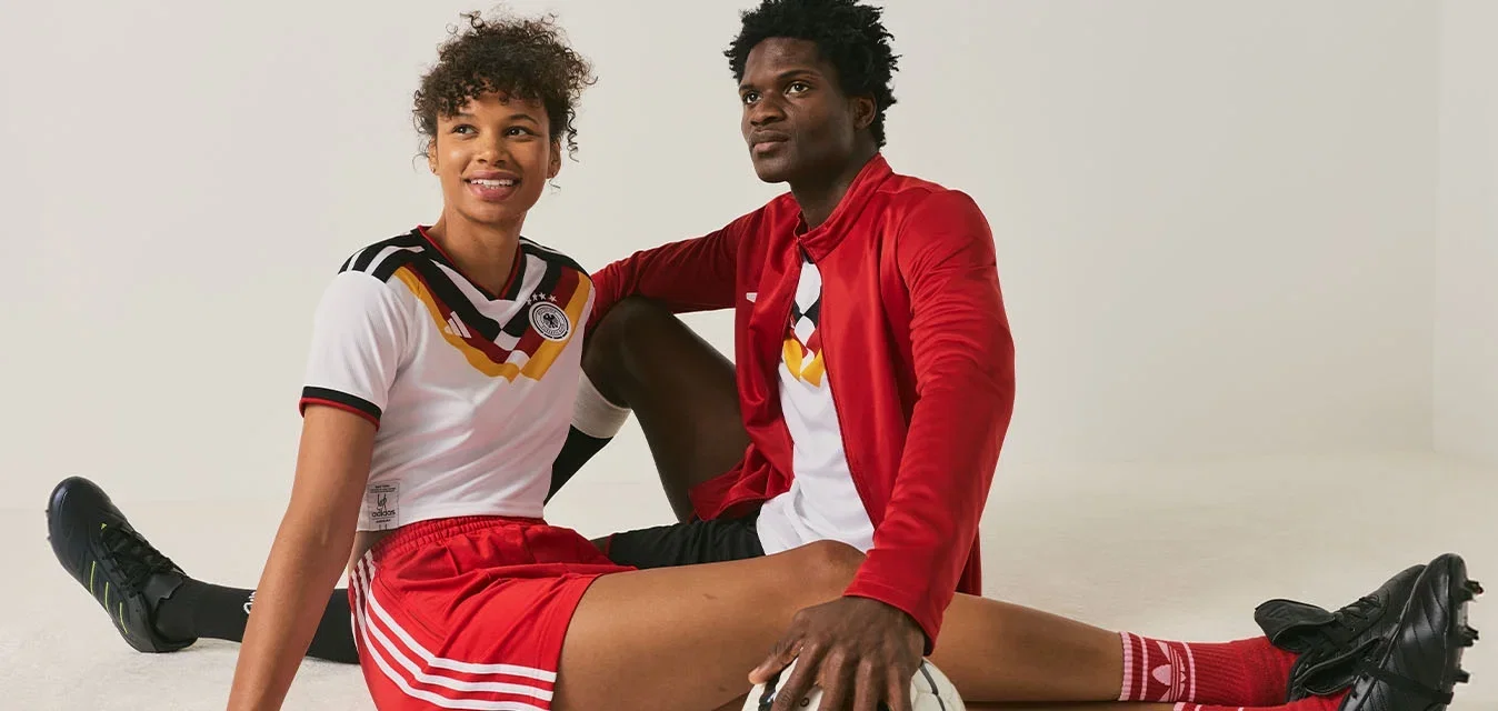 Dos jóvenes jugadores de fútbol con uniformes de equipo sentados en el suelo, uno sosteniendo un balón de fútbol, sobre un fondo blanco liso.