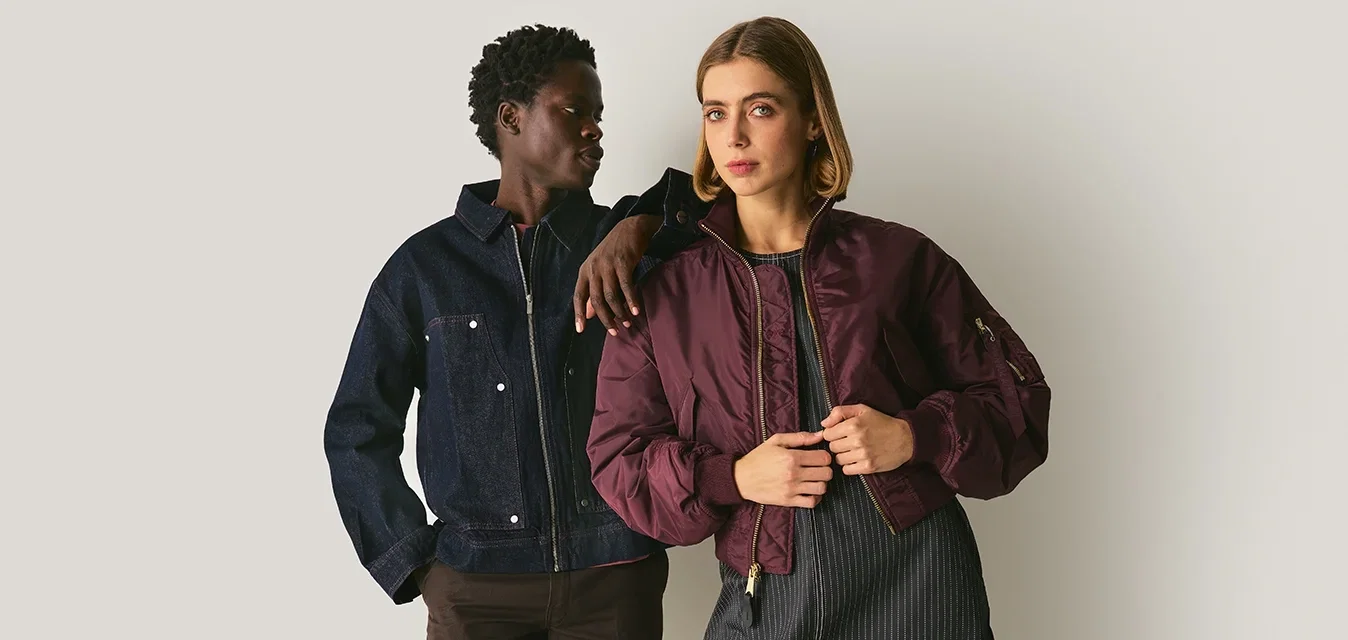 Dos modelos posan juntas. Una lleva una chaqueta de mezclilla oscura y pantalones marrones; la otra viste una chaqueta bomber burdeos sobre un vestido a rayas.