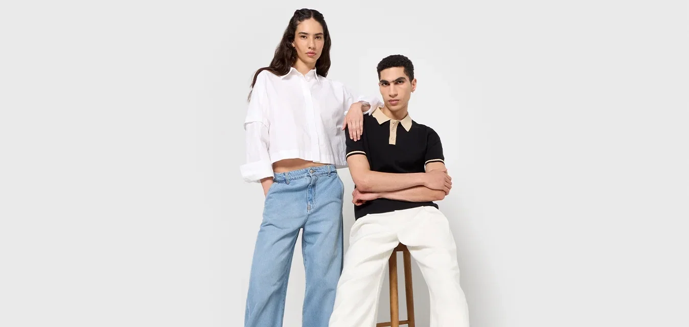 Twee modellen staan naast elkaar. De ene draagt een wit crop-top shirt en een blauwe wijde spijkerbroek; de andere draagt een zwart polo en witte broek.