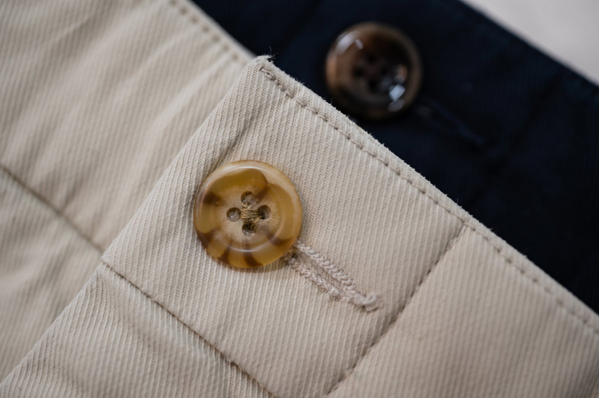 A close up of the button area of the Bills Khakis Ilsand Twill khaki pants, with the navy pair semi out of focus in the background