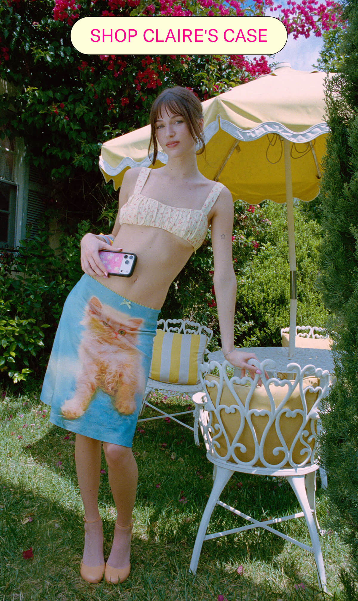 Claire standing in a garden holding the Claire Drake x Wildflower phone case, wearing a kitten-print skirt
