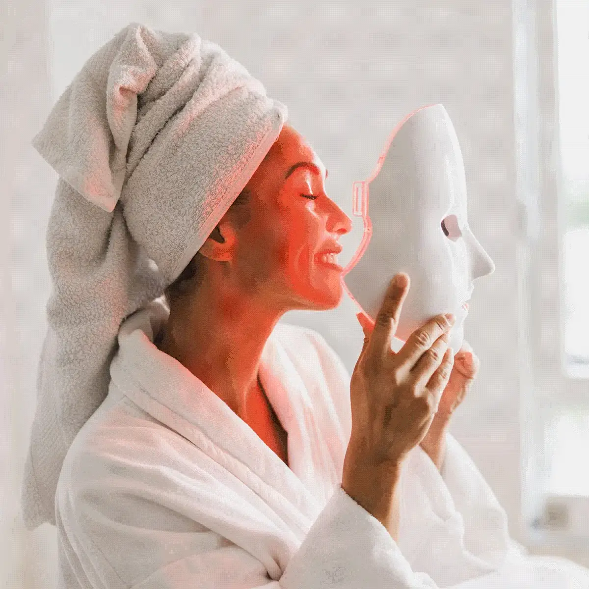 Woman using white facial steaming mask device, wearing towel wrap, in spa skincare routine