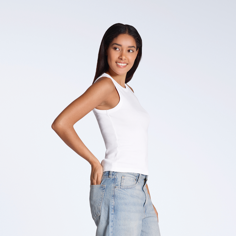 A woman smiling with her hand in her back pocket, modelling a white, organic cotton tank top.