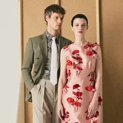 Homme en blazer vert olive et pantalon beige debout à côté d’une femme en robe fleurie rose sur un fond beige minimaliste.
