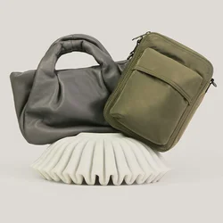 Zwei Handtaschen, eine graue Handtasche aus weichem Leder und eine olivgrüne rechteckige Umhängetasche, auf einem weißen, strukturierten Ständer präsentiert.