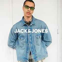Mann mit grüner Sonnenbrille, hellblauer Jeansjacke über einem karierten Hemd, blauer Jeanshose und schwarzen Schuhen steht mit den Händen hinter dem Rücken an einer weißen Wand.