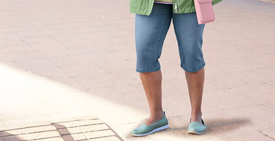 Ein Model trägt eine eucalyptus farbene Übergangsjacke, darunter ein weiß-orchidee-geringeltes Shirt und eine blue-bleached Caprijeans