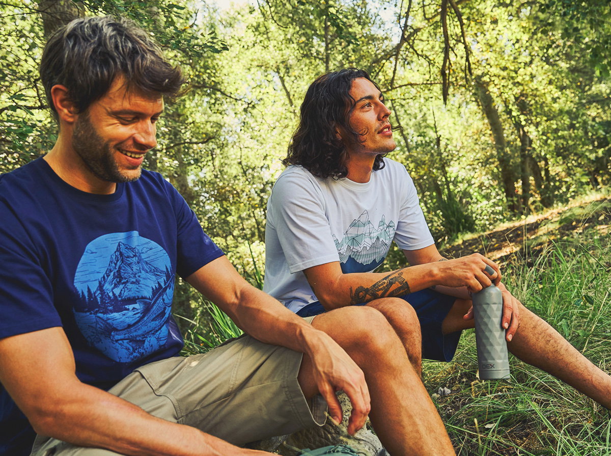 Two people sitting on grass in a leafy forest with sunlight filtering through the trees, wearing t-shirts and shorts, resting during an outdoor hike; one person holds a reusable water bottle while both sit among green grass on a gentle slope.