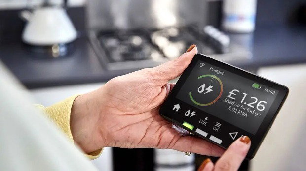 A person standing by a gas hob in a kitchen holds a smart meter, which has different figures and measurements displayed on its screen.