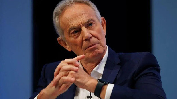 Tony Blair folds his hands together as he looks slightly to the side. He is wearing a dark blue blazer with a white shirt.