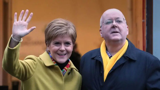 Nicola Sturgeon and Peter Murrell stand outside the entrance to a building. Sturgeon, wearing a light green coat and colourful scarf, raises a hand in a wave, while Murrell wears a dark coat and bright yellow scarf and looks ahead. The open doorway behind them reveals warm indoor lighting, indicating they are just outside.