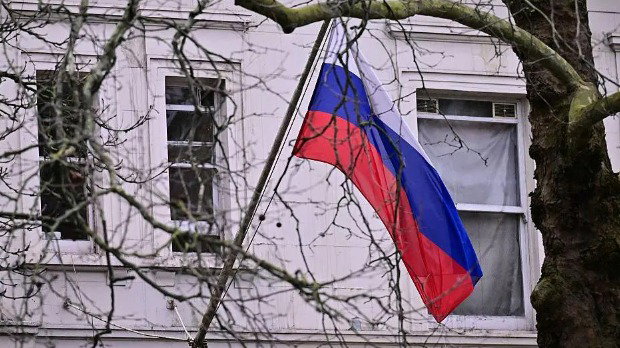 A white, blue and red Russian flag hangs from a pole in front of a white building and behind a tree that has no leaves on its branches.