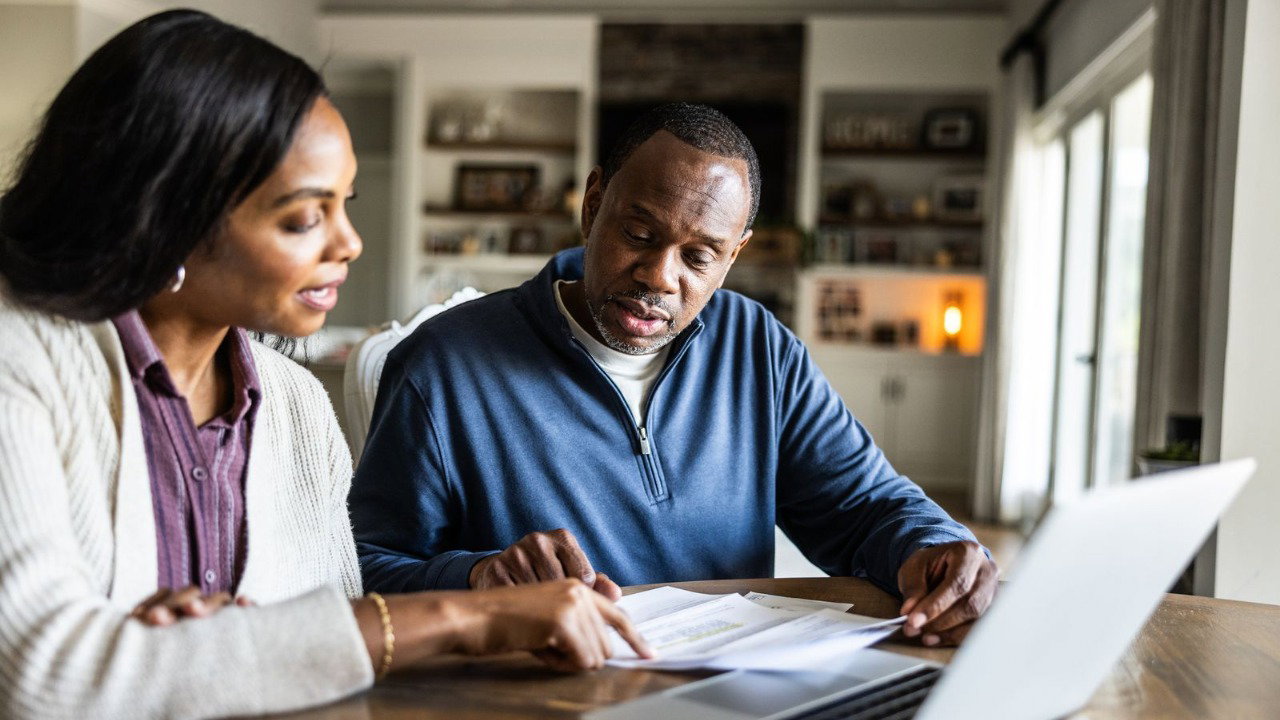 A man and a woman look at pieces of paper, that are laid out on a dark wooden table in front of a silver laptop. They are seated in a sitting room of a house.