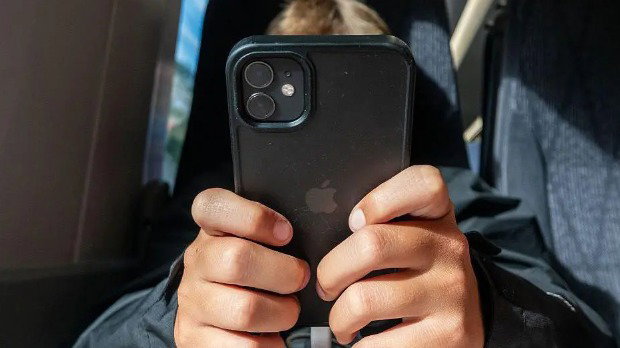 A persons face is obscured as they use a phone on a train. They are sat on dark blue seats and the phone is black.