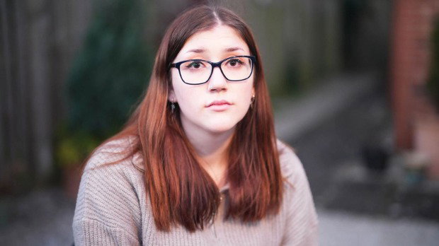 A close-up portrait of Laura Kenny, facing the camera directly. She has long brown hair that falls past her shoulders and she is wearing dark-framed glasses. Her expression is calm and neutral.
