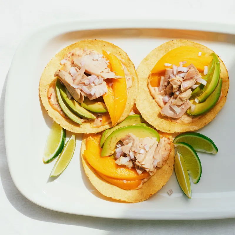 Three tostadas with canned tuna, sliced mangoes, sliced avocados, shallots, and spicy mayo on a platter served with lime wedges.
