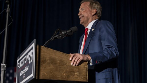 Republican Senate nominee Ken Paxton stands at a podium, speaking to supporters at a watch party. He wears a blue suit and a red tie. 
