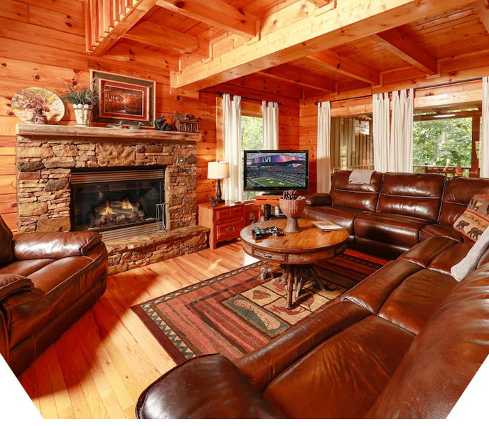 Rustic log cabin family room with leather sectional, stone fireplace, wood beams, round coffee table, and TV.