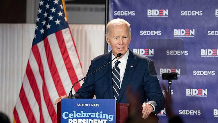Joe Biden speaks at an event for South Carolina Democrats in February 2026. He stands at a podium that is fitted with a sign 'Celebrating President Biden'. An American flag hangs in the background. He wears a dark suit, striped tie and white shirt.
