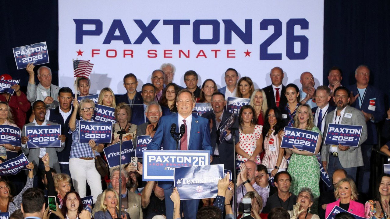 Ken Paxton smiles from a podium at a rally. He wears a blue suit, white shirt and red tie.