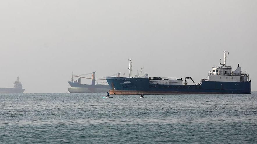 Several large cargo and container ships are anchored in hazy conditions at sea near the Strait of Hormuz, with two small figures on paddleboards or watercraft floating in the foreground.