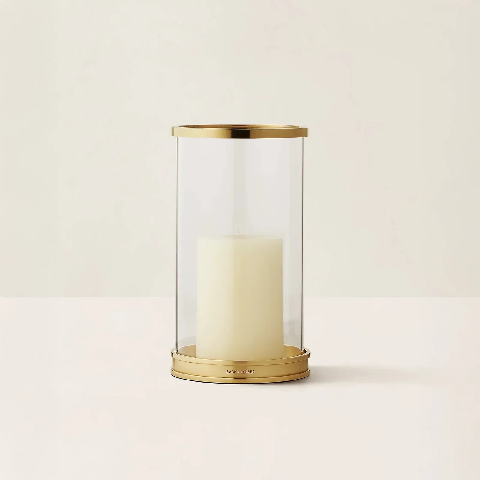 Clear glass hurricane candle holder with gold rim and base holding an unlit ivory pillar candle on pale background