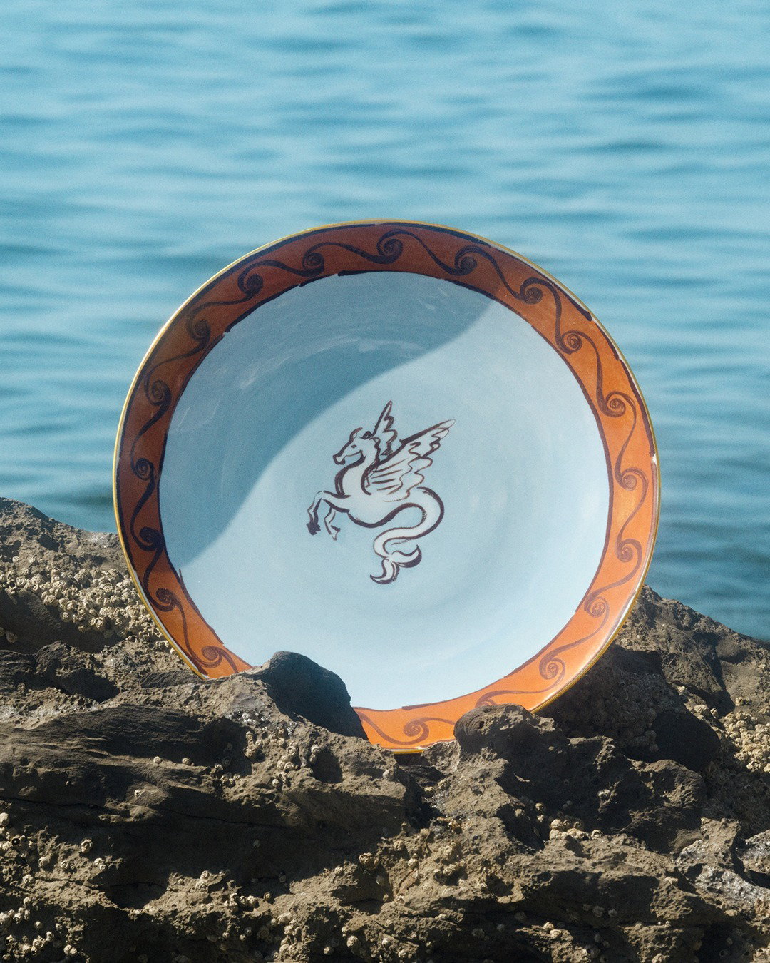 Ceramic plate with orange decorative rim and central winged horse (Pegasus) motif, propped on rocky shore with sea background