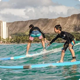 Two young boys smiling and surfing on long blue surfboards in the ocean with mountains in the background.