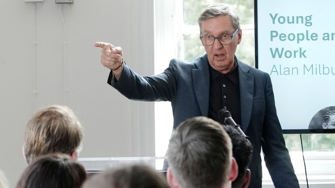 Alan Milburn, who has grey hair and wears a dark grey suit jacket with black polo shirt, points off to the side as he speaks to young people. His name is on a projector behind him.