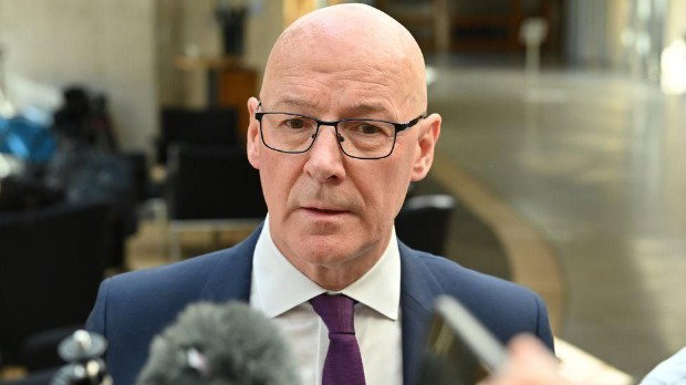 John Swinney, who is bald with glasses, speaks into microphones. He is wearing a blue business suit with a purple tie.