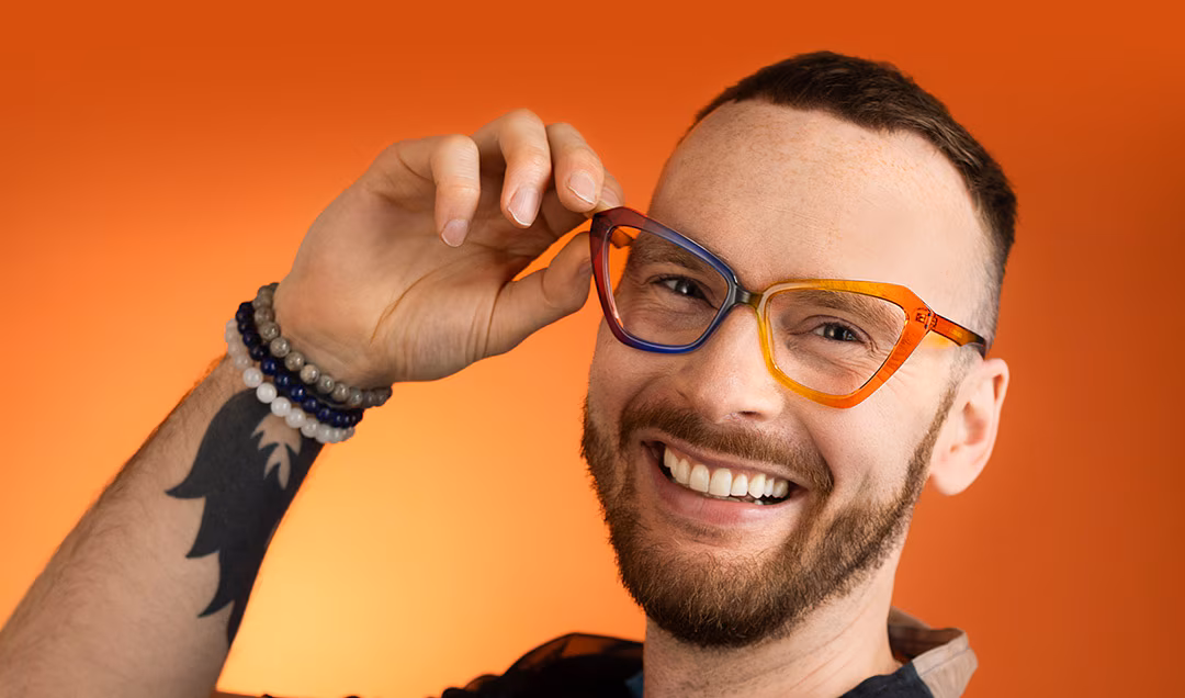 Smiling man with a beard and arm tattoo, holding up The Collection Notice multi-coloured cat-eye glasses against an orange background