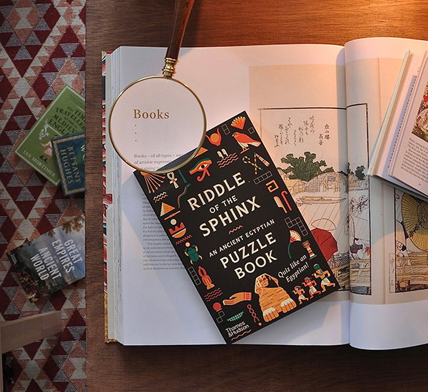Large illustrated book open on a desk with a magnifying glass highlighting the word “Books” on the open page and a smaller riddle book next to it. - Shop Now.