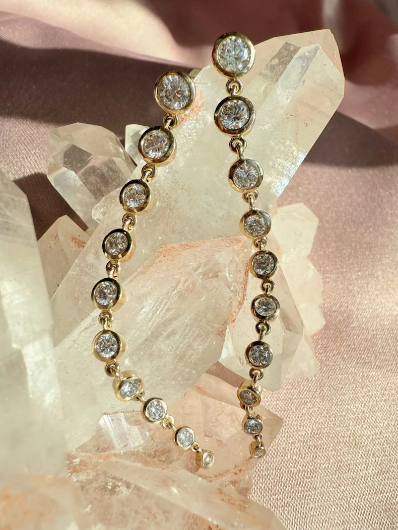 Dangle diamond earrings draped over quartz crystals on a blush satin surface.
